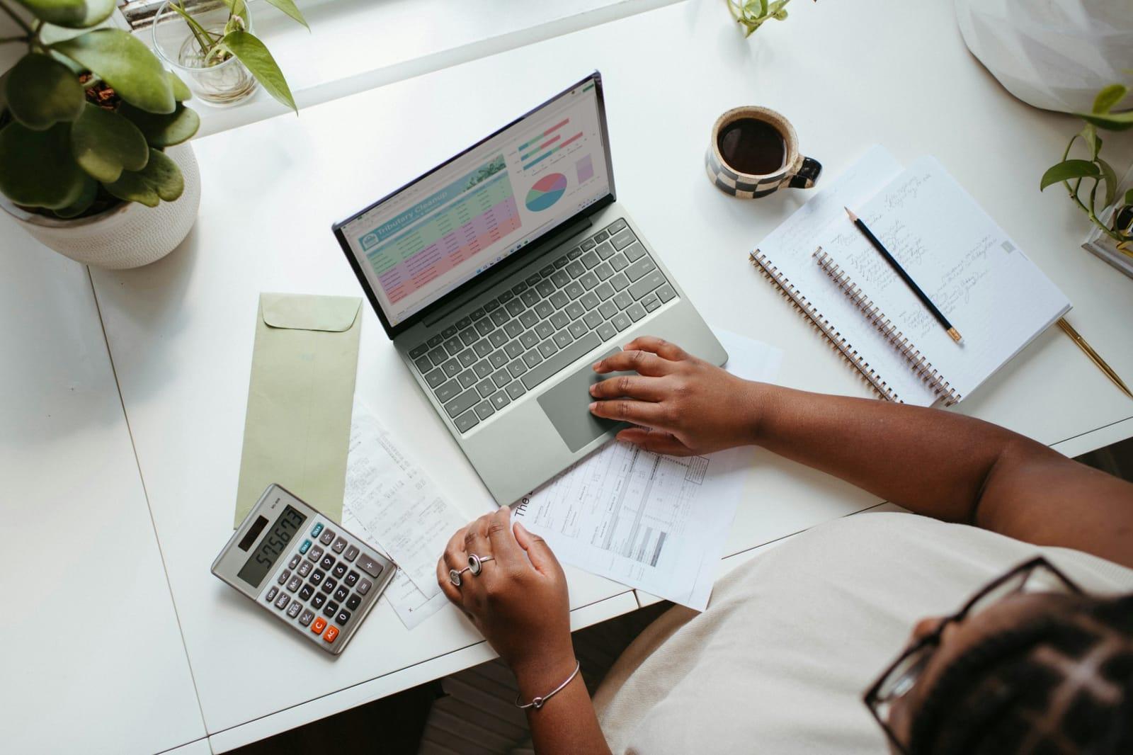 A person at a desk working on a laptop spreadsheet beside a calculator, receipts and a notebook